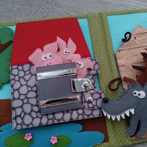 May include: A felt quiet book page featuring a scene from the Three Little Pigs. The page shows a red-roofed stone house with three pink pigs, a wooden house, a gray wolf, a tree, and a pond with lily pads.