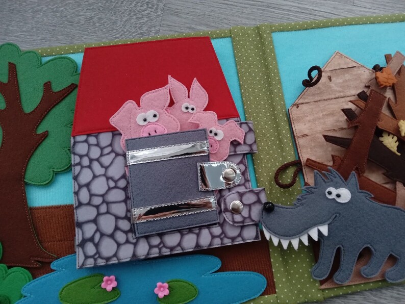 May include: A felt quiet book page featuring a scene from the Three Little Pigs. The page shows a red-roofed stone house with three pink pigs, a wooden house, a gray wolf, a tree, and a pond with lily pads.
