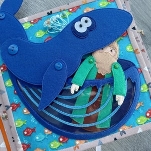 May include: A handmade felt quiet book page featuring a blue whale with a person inside. The whale is attached with buttons. The background is a colorful fabric with fish. The book has snap closures and a variety of textures.