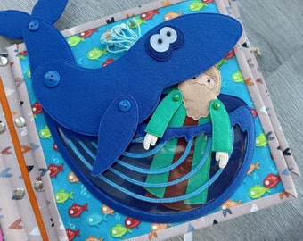 May include: A handmade felt quiet book page featuring a blue whale with a person inside. The whale is attached with buttons. The background is a colorful fabric with fish. The book has snap closures and a variety of textures.