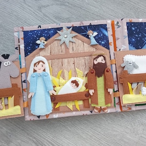 Op de afbeelding: Een handgemaakt kerststalboek van vilt met meerdere pagina's. De pagina's tonen het kerstverhaal met vilten figuren van Maria, Jozef, baby Jezus, engelen en dieren. De achtergrond heeft een sterrenhemel en een stal.