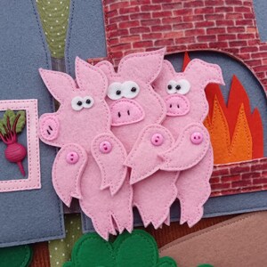 May include: A felt craft depicting the Three Little Pigs story. Three pink pigs with button eyes and noses stand in front of a brick chimney with orange felt flames. A window shows a pink-framed radish.