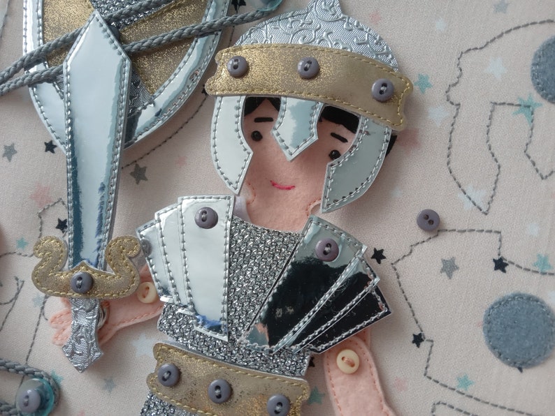 May include: A fabric applique of a knight in silver and gold armor, holding a sword and shield. The knight has a smiling face and is adorned with buttons. The background is a light pink fabric with star and constellation patterns.
