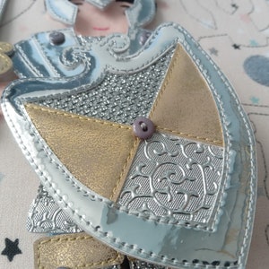 May include: A close-up of a decorative applique featuring a knight's armor design. The applique has a silver, gold, and gray color scheme with a shiny, metallic finish. The shield is detailed with embossed patterns and a purple button.