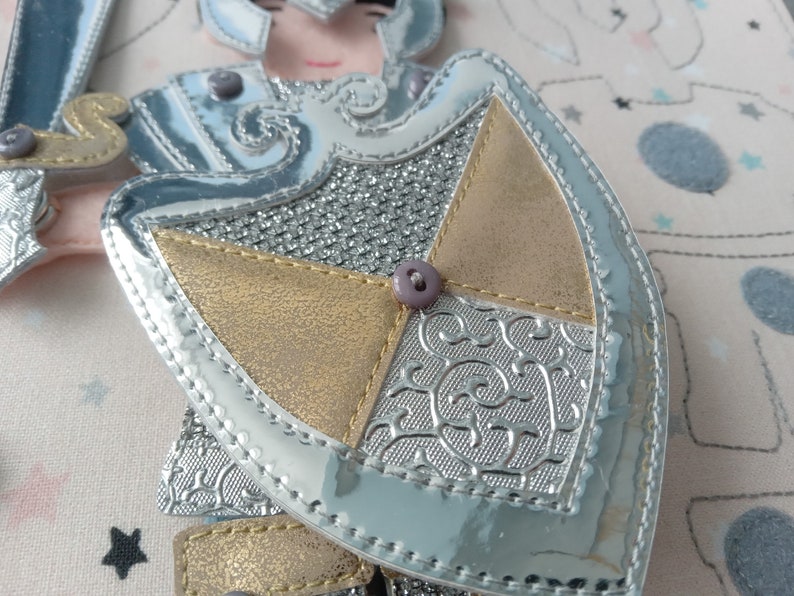 May include: A close-up of a decorative applique featuring a knight's armor design. The applique has a silver, gold, and gray color scheme with a shiny, metallic finish. The shield is detailed with embossed patterns and a purple button.