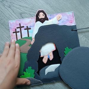 Op de afbeelding: Een vilten paas-thema rustig boek pagina. De scène toont Jezus die uit een graf komt, met drie kruisen op een heuvel op de achtergrond. De pagina heeft een verwijderbare steen en is gemaakt van verschillende kleuren.