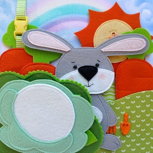 May include: A felt bunny with grey fur, a black nose, and pink cheeks, surrounded by felt carrots, a sun, and a flower. The background features a rainbow and green patterned fabric. A yellow buckle is visible.