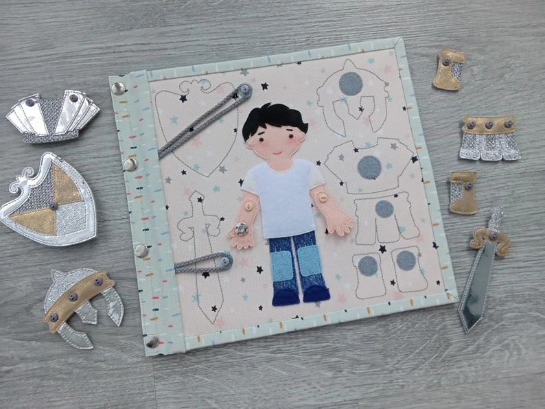 May include: A fabric activity board with a knight theme. The board features a felt figure of a boy with removable clothing pieces, including a helmet, armor, and sword. Additional felt knight accessories are scattered around the board.