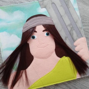 Puede incluir: Muñeca de fieltro que representa una figura con cabello castaño largo, ojos azules y una diadema gris. La muñeca sostiene un objeto gris y plateado y lleva una prenda amarilla. El fondo presenta un cielo con nubes.