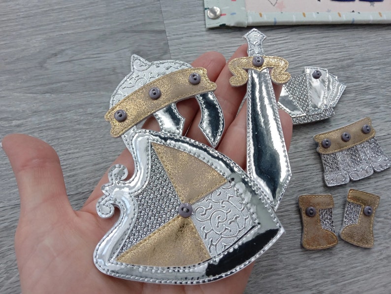May include: A set of silver and gold knight costume pieces, including a helmet, shield, sword, and boots. The pieces are made of metallic fabric and feature decorative details and button accents. The items are designed for crafting or costume use.