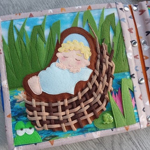 May include: A handmade fabric quiet book page featuring a sleeping baby in a woven basket. The scene includes green felt grass, a blue water background, and a green frog. The book has a blue button closure and a fish pattern.