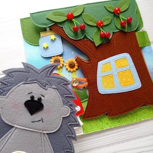 May include: A colourful felt activity book featuring a grey hedgehog, a brown tree with a window, and a green leafy top with red cherries. The book also includes a blue jar with yellow stars and two yellow sunflowers.