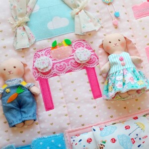 May include: A handmade, quilted play mat with a house theme. Features include a pink table with lace trim, felt curtains, and two bunny dolls. The dolls wear blue overalls and a floral dress. The mat is decorated with gold hearts.