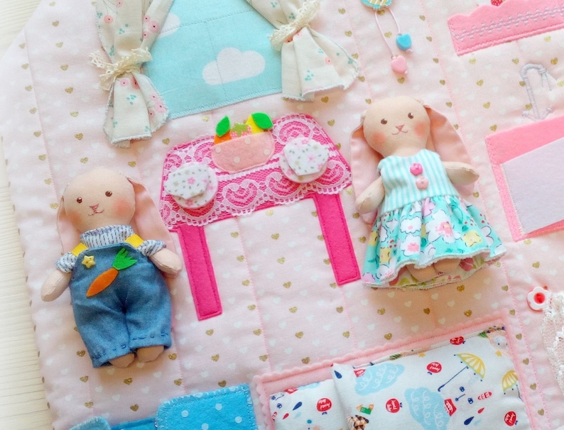 May include: A handmade, quilted play mat with a house theme. Features include a pink table with lace trim, felt curtains, and two bunny dolls. The dolls wear blue overalls and a floral dress. The mat is decorated with gold hearts.