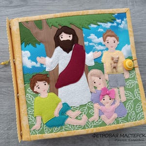 Op de afbeelding: Een handgemaakt vilten boek met een religieus thema. De omslag toont een scène met een figuur in witte gewaden en een rode sjerp, omringd door kinderen. De achtergrond omvat een boom, blauwe lucht en groen gras. Het boek is gebonden met metalen ringen.