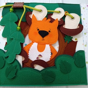 May include: A felt activity book featuring an orange fox with a white bib, surrounded by green trees, brown tree trunks, and brown mushrooms. The book has a green border and a green and brown cord with a wooden bead. The background is white with colorful dots.