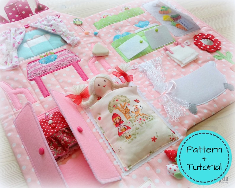 May include: A pink and white polka-dotted fabric activity mat with various interactive elements. Features include a doll, a table, a bed, a closet with a dress, and other felt and fabric accessories. The mat has a variety of textures and colors.