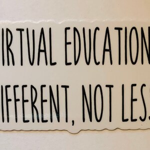 May include: A white sticker with black text that reads "VIRTUAL EDUCATION DIFFERENT, NOT LESS." The sticker has a slightly irregular shape and is likely designed for laptops, notebooks, or other surfaces.