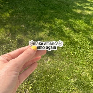 May include: White sticker with black text that reads "Make America Emo Again" with two black hand drawn rock and roll hand gestures on either side of the text.