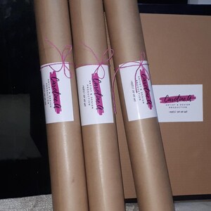Pode incluir: Tr&ecirc;s tubos de papel castanho com um cord&atilde;o rosa amarrado &agrave; volta. Cada tubo tem um r&oacute;tulo branco com o texto "Caroline A" e "Artist & Design Production" em preto.