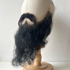 Puede incluir: Una peluca de barba negra completa sobre una cabeza de maniquí. La peluca está diseñada con un diseño largo y fluido. La cabeza del maniquí es de color beige claro y está montada sobre un soporte de madera. El fondo es blanco liso.