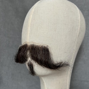 May include: A dark brown mustache and goatee attached to a flesh-toned lace base, displayed on a mannequin head. The facial hair is styled with a full mustache and a small goatee. The mannequin head is covered in a light beige fabric.