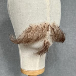 May include: A brown and light brown realistic mustache and goatee attached to a fabric head form. The facial hair has a natural appearance. The head form is mounted on a wooden stand. The head form is on a wooden stand.