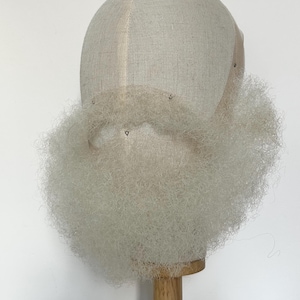 Peut inclure: Une barbe blanche, bouclée et texturée, composée de fines fibres synthétiques fixées sur une tête de mannequin en tissu beige. La barbe est fournie et volumineuse, s'étendant du menton et de la mâchoire, fixée par une base en maille et des épingles.
