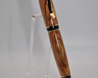 May include: A wooden pen with a gold trim and black accents. The pen is made of olive wood and has a unique grain pattern. It is displayed on a clear acrylic stand.