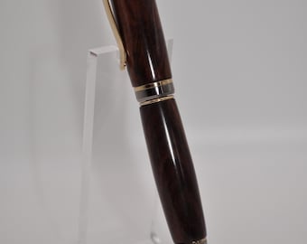 May include: A brown wooden pen with gold accents and a silver band. The pen is on a clear acrylic stand.