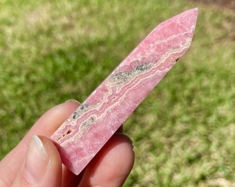 May include: A pink and white rhodochrosite crystal point. The crystal has a layered appearance with bands of pink and white. The top of the crystal is a pointed shape. The crystal is held in a hand against a background of green grass and trees.