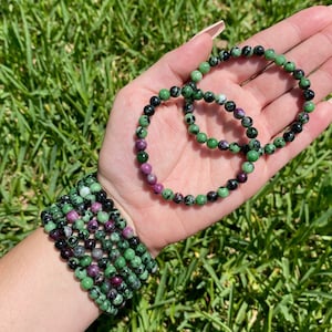 May include: A set of green, black and purple gemstone bracelets. The bracelets are made of small, round beads and are stacked on a person's wrist.