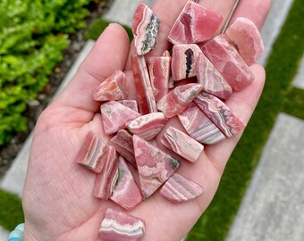 May include: A collection of pink and white striped, triangular shaped, polished gemstones. The stones are arranged in a pile on a person's hand.