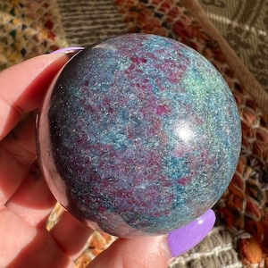 Peut inclure: Une sphère polie de zoïsite rubis, également connue sous le nom d'anyolite, avec une couleur vert foncé, rouge et bleu. La sphère est lisse et a une finition brillante.