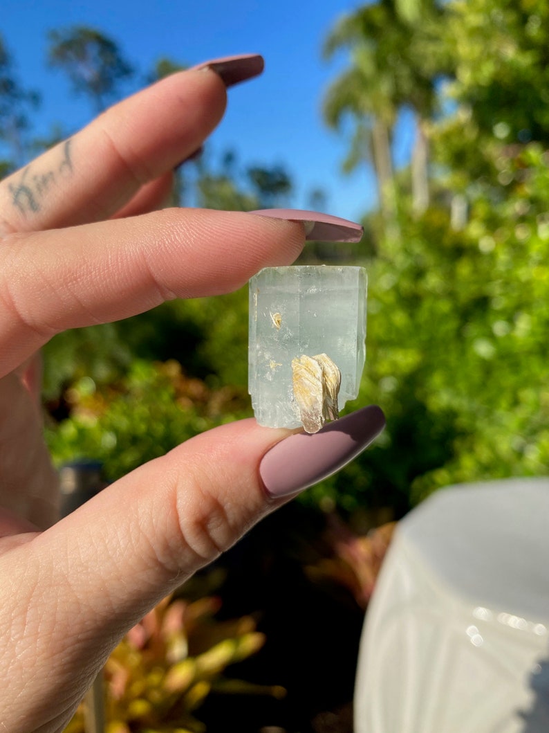 May include: A light blue aquamarine crystal with a clear, glassy texture. The crystal has a natural, rough shape and is held in a hand.