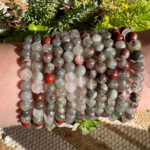 Pu&ograve; includere: Un set di braccialetti realizzati con perline di pietre preziose verdi, rosse e bianche. I braccialetti sono impilati sul polso di una persona.