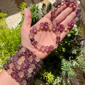 Puede incluir: Un conjunto de tres pulseras hechas con cuentas de fluorita p&uacute;rpura y amarilla. Las pulseras est&aacute;n apiladas en la mu&ntilde;eca de una persona.