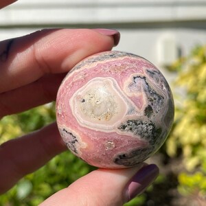 May include: A pink and black sphere of polished gemstone with swirling patterns. The sphere is smooth and has a high gloss finish.