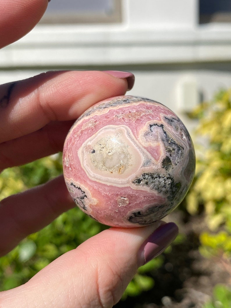 May include: A pink and black sphere of polished gemstone with swirling patterns. The sphere is smooth and has a high gloss finish.