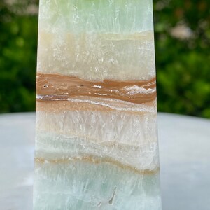 May include: A green, white, and brown layered obelisk crystal. The crystal has a smooth, polished surface and is a natural stone.