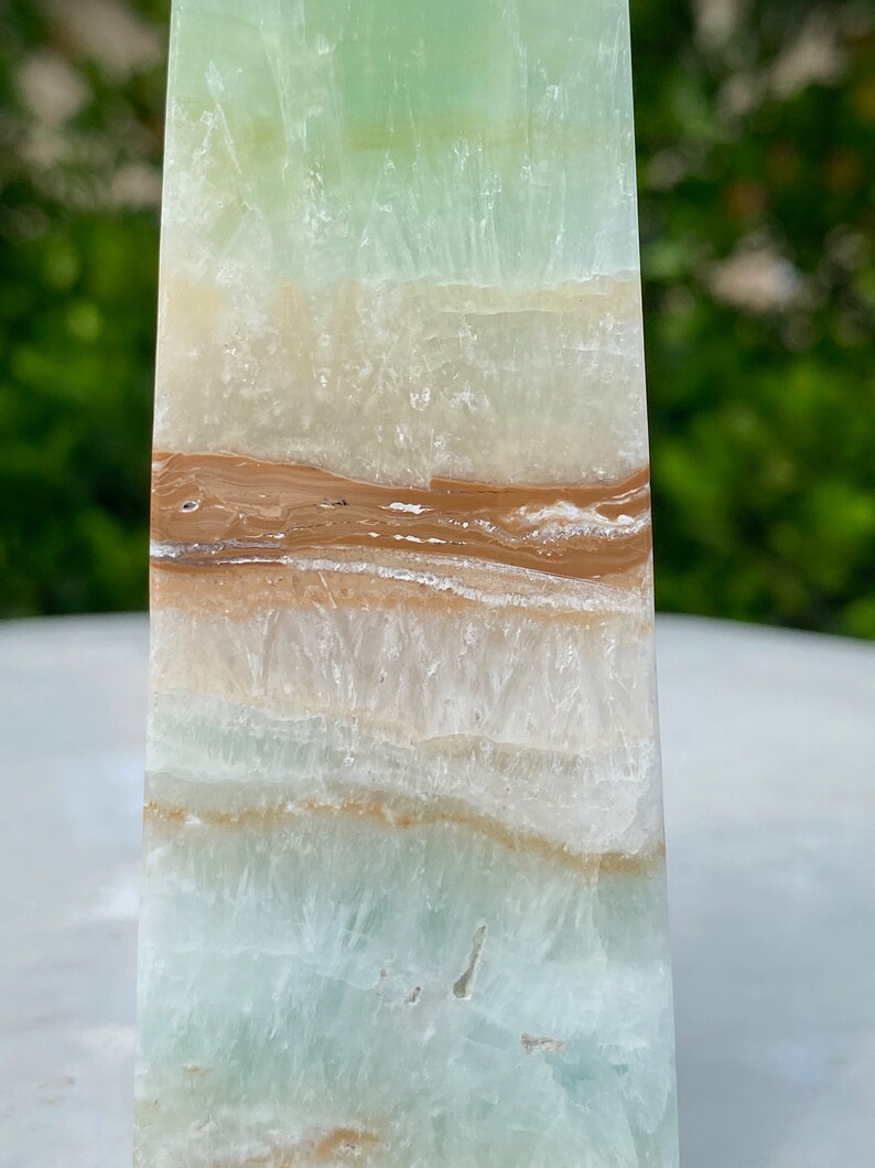May include: A green, white, and brown layered obelisk crystal. The crystal has a smooth, polished surface and is a natural stone.