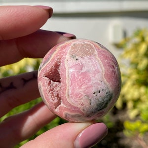 May include: Pink and white banded sphere of polished pink calcite, a natural crystal with a unique pattern. The sphere is smooth and has a slightly rough texture.