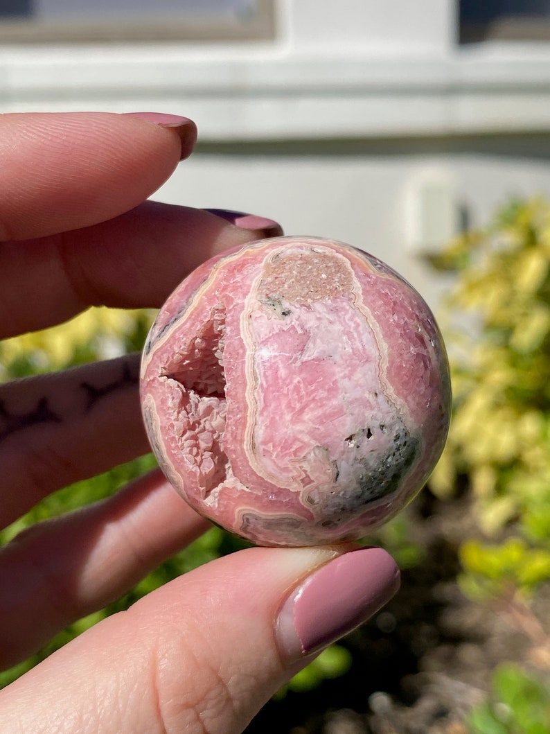 May include: Pink and white banded sphere of polished pink calcite, a natural crystal with a unique pattern. The sphere is smooth and has a slightly rough texture.