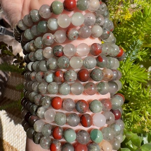 Pu&ograve; includere: Un set di braccialetti realizzati con perline di pietre preziose verdi, rosse e bianche. I braccialetti sono sovrapposti sul polso di una persona.