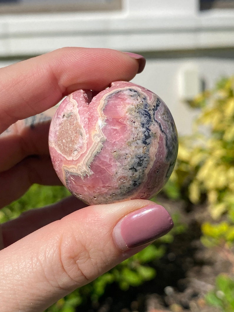 May include: A pink and gray sphere of polished pink manganese, also known as rhodochrosite, with a swirling pattern of pink, white, and gray.