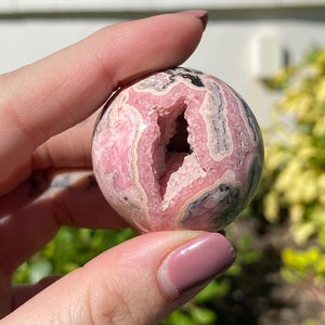 May include: A pink and gray gemstone sphere with a large, natural opening. The sphere is smooth and polished, with a variety of pink and gray bands.
