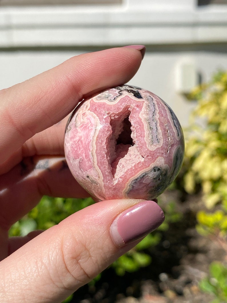 May include: A pink and gray gemstone sphere with a large, natural opening. The sphere is smooth and polished, with a variety of pink and gray bands.