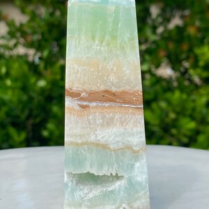 May include: A green, white, and brown banded obelisk crystal point. The crystal is polished and has a smooth, glassy surface.