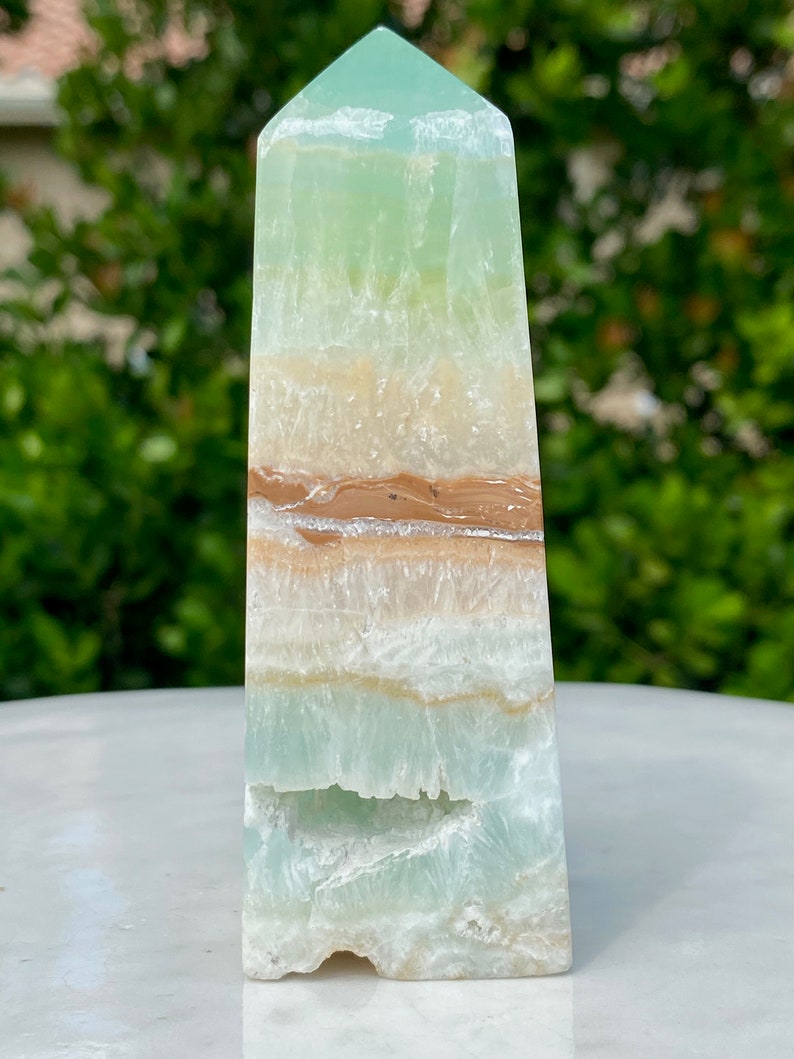 May include: A green, white, and brown banded obelisk crystal point. The crystal is polished and has a smooth, glassy surface.