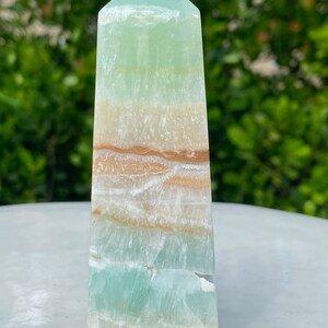 May include: A green, white, and brown banded onyx obelisk crystal point. The crystal is polished and has a smooth, glassy finish.
